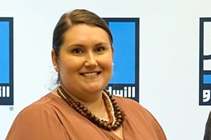 Smiling woman with brown hair wearing a peach blouse and beaded necklace, standing in front of a blue logo backdrop.