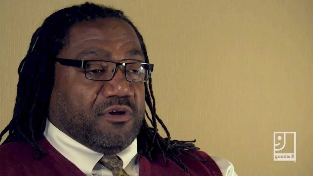 Man with dreadlocks and glasses speaks, wearing a burgundy sweater over a white shirt and tie, against a beige wall.