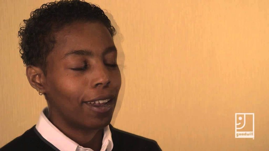 Person with short curly hair, eyes closed, smiling softly against a beige wall; Goodwill logo bottom right.