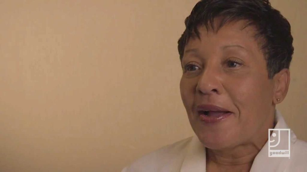 Close-up of a smiling woman with short dark hair wearing a light jacket, against a beige background, with a Goodwill logo.