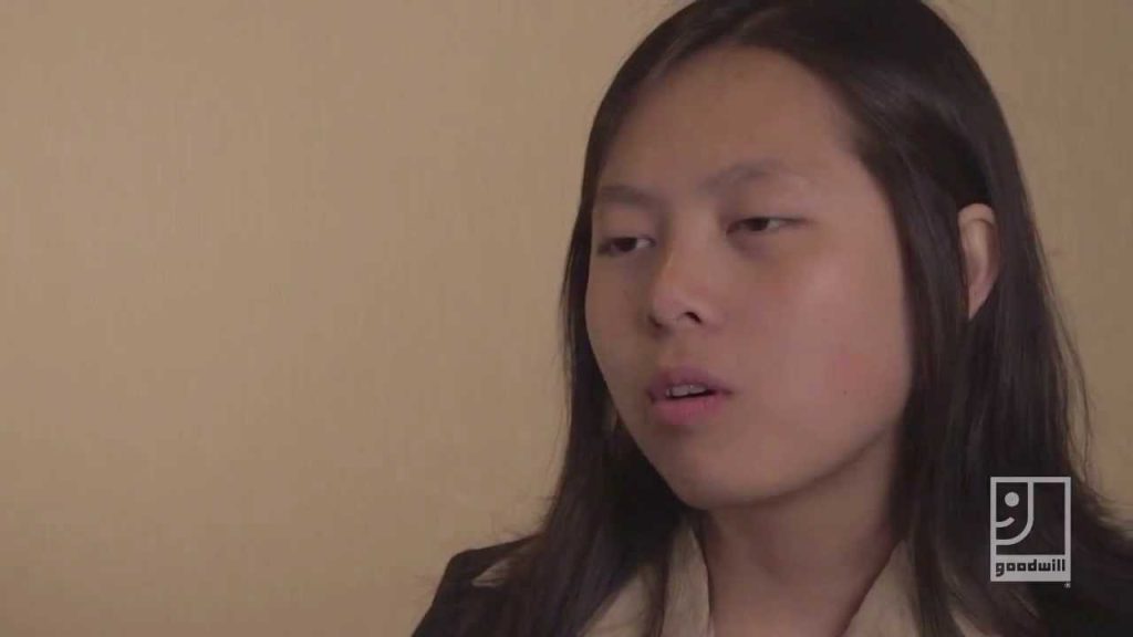 Young woman with long dark hair, looking slightly to the side, against a beige wall.