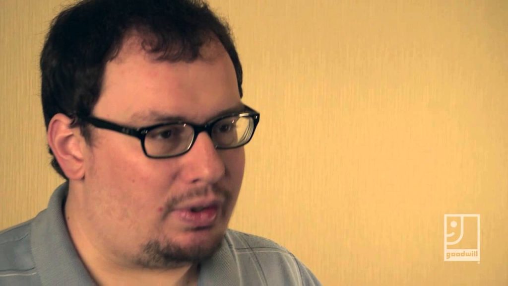 Man with dark hair and black glasses, wearing a gray polo, looking to the right; beige wall and a goodwill logo.