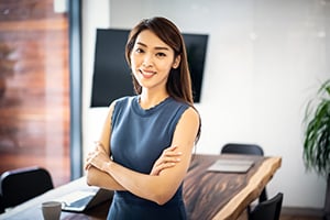 Confident professional woman in a blue sleeveless top stands with arms crossed in a modern conference room.