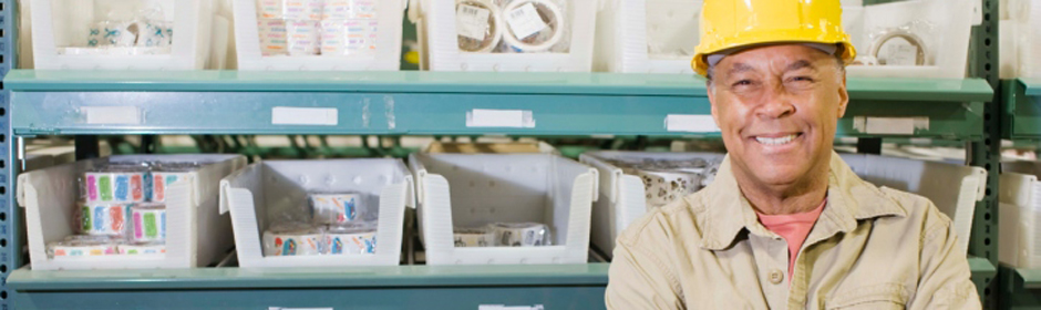 Smiling worker in a yellow hard hat standing among labeled bins on metal warehouse shelves.