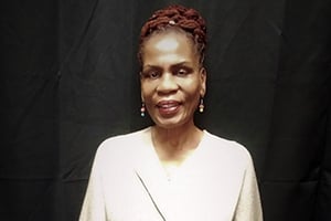 Portrait of a smiling woman with braided hair wearing a light beige blazer against a dark backdrop.