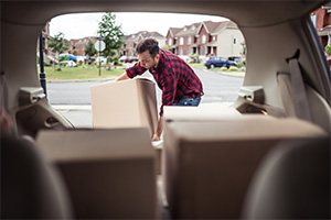 Man unloading moving boxes from a car trunk in a suburban driveway.