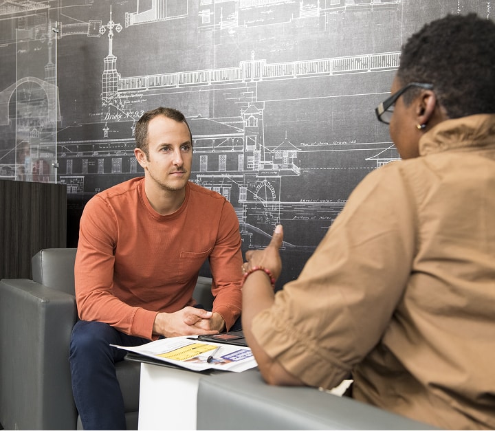 Two coworkers in a casual office lounge discuss documents with a large blueprint wall behind them.
