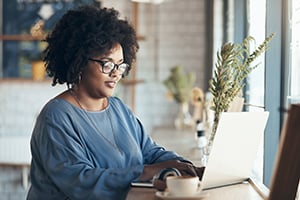 Woman with curly hair and glasses working on a laptop at a cafe desk.