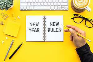 Open notebook on a bright yellow desk shows 'New Year New Rules' with a hand writing in it, surrounded by office supplies.