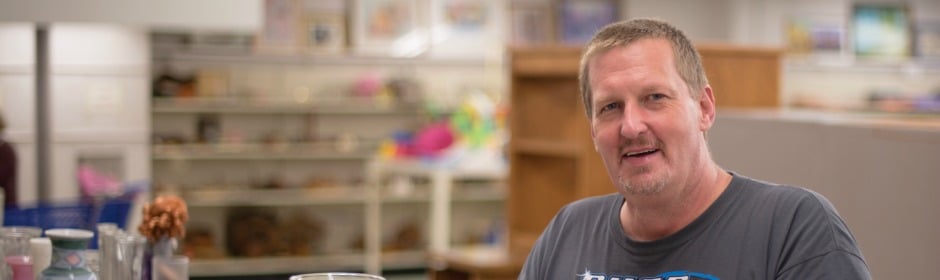 Smiling man in a casual T-shirt stands in a shop with shelves and glassware in the background.