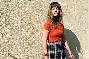 Young woman with blunt bangs wearing an orange top and plaid skirt stands against a textured beige wall.
