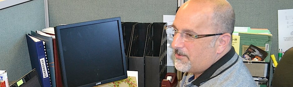 Man with glasses sits at a cubicle desk with a computer monitor and organized folders nearby.