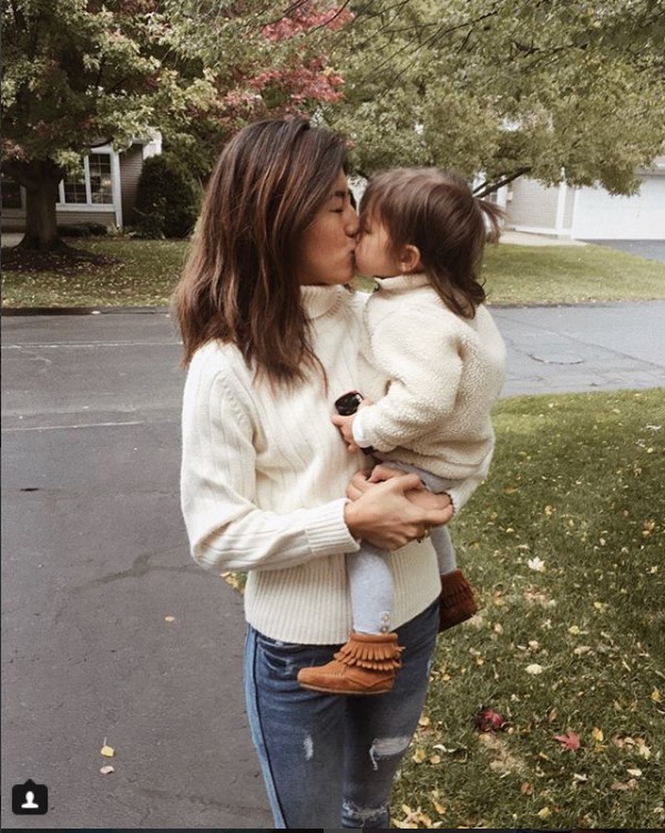 Mother in a cream knit sweater holds and kisses her toddler outdoors on a fall day.