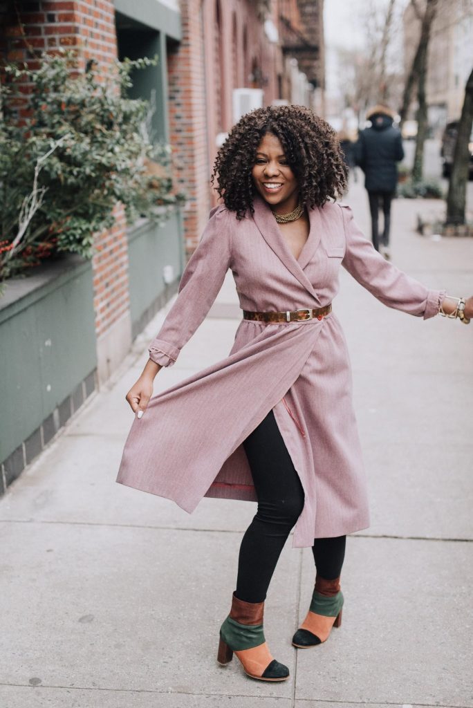 Smiling woman in a pink belted trench coat walking on a city sidewalk.