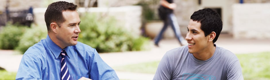 Two men seated outdoors in conversation; one wears a blue dress shirt and striped tie, the other a gray t-shirt.