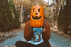 Person wearing a carved pumpkin mask sits cross-legged on a leaf-covered path in autumn.