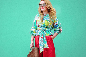 Stylish woman in a tropical-print blouse and red pants stands against a teal background with a woven handbag.