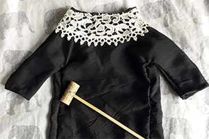 Black doll-sized dress with an intricate white lace collar laid on light fabric.