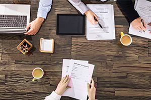 Top-down view of a business meeting around a wooden table with laptops, tablets, documents, and coffee cups.