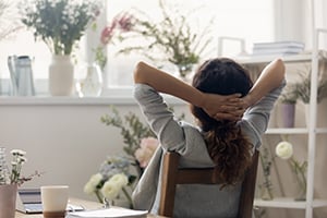 Person with long hair sits relaxed at a bright home office, hands behind head, with plants and a coffee mug nearby.