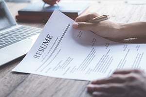 Close-up of a hand signing a resume on a wooden desk, with a laptop blurred in the background.