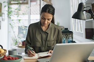 Woman writing in a bright home office, working at a laptop with a notebook beside her.