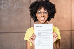 Smiling woman with curly hair and glasses holds a resume toward the camera.
