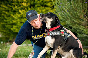 Trainer kneels beside a black-and-white working dog wearing a Julius-K9 harness outdoors.