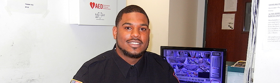 Smiling security guard in a dark uniform sits beside a multi-camera security monitor.