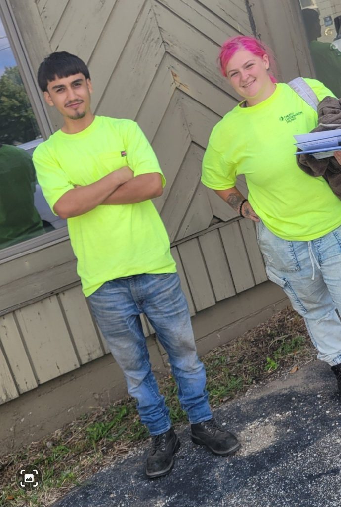 Two workers in neon shirts pose outside a wooden building; left man crosses arms, right person with pink hair holds papers.