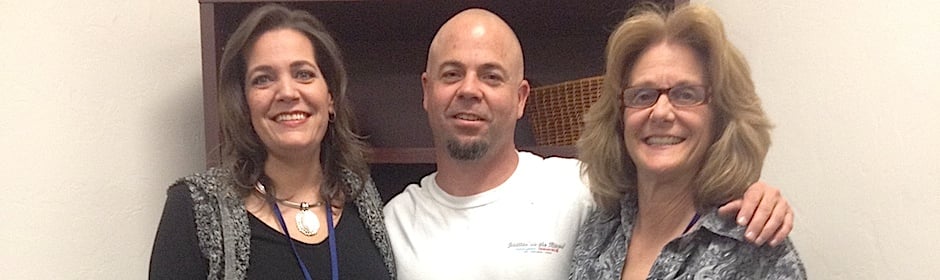Three adults smiling for a group photo indoors, two women on either side of a bald man wearing a white t-shirt.