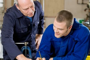 Two technicians in blue uniforms collaborate over a workshop project, with an older mentor guiding the younger trainee.
