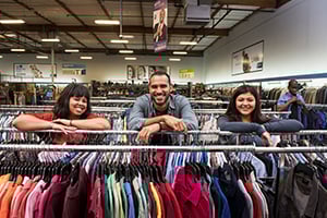 Three shoppers lean on a clothing rack, smiling in a busy store filled with colorful clothes.