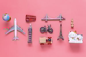 Miniature travel items on a pink background: airplane, globe, bus, bike, bridge, Big Ben, Eiffel Tower, and a sofa with a cat.