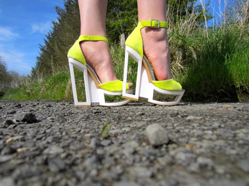 Bright lime-green high-heeled platform sandals with white chunky heels on a gravel path outdoors.