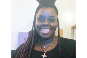 Smiling woman with long box braids, wearing a black top and a pendant necklace.