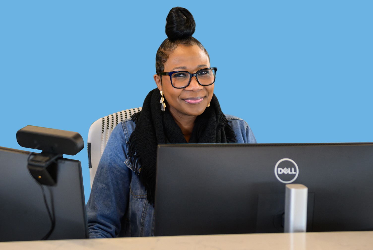 Smiling woman with black-framed glasses sits at a desk with a Dell monitor against a blue background.