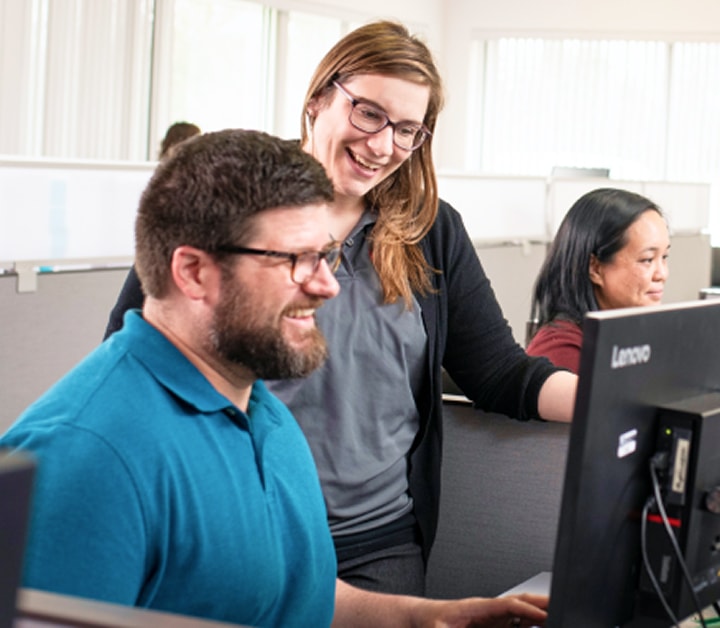 Bearded man in a blue polo works at a Lenovo computer while a smiling woman leans over his shoulder in a bright open office.