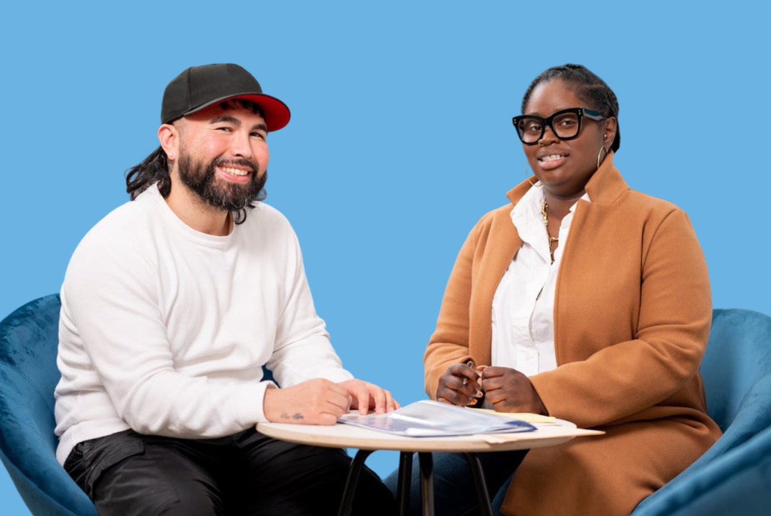 Two professionals sit on blue chairs at a round table, reviewing documents against a solid blue background.