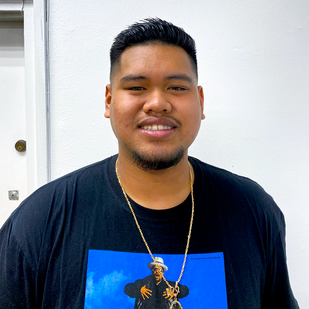 Portrait of a smiling young man with short black hair and a goatee, wearing a black T-shirt and a gold chain.