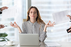 Businesswoman meditating with laptop at a desk as papers float around in a modern office.