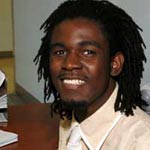 Smiling man with dark skin and shoulder-length dreadlocks, wearing a light-colored shirt and tie.