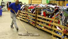 A worker mops the floor in a thrift store with clothing racks and wooden barriers nearby.
