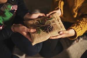 People presenting a small burlap-wrapped gift tied with twine and a pinecone