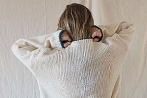 Back view of a person wearing an oversized cream knit sweater, hands near their head.