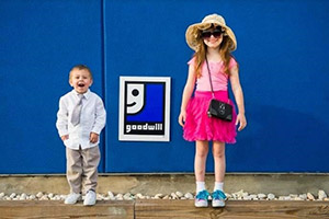 Two kids stand on a wooden platform in front of a blue wall with a Goodwill sign; boy in a white shirt and beige pants, girl in a pink dress with a hat.
