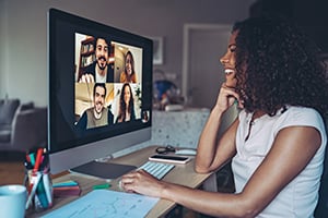 Woman at a home desk participates in a video conference with multiple participants on a computer screen.