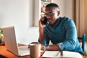 Man in a blue denim shirt wears glasses while talking on a cellphone at a wooden desk with laptop, mug, and notebook.