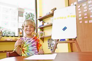 Smiling child in a tie-dye shirt displays artwork at a classroom table.
