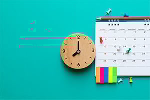 Wooden clock mounted on teal wall beside a calendar with colorful sticky notes.
